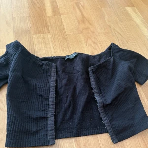Svart offshoulder topp från Bershka - Säljer en svart offshoulder topp från Bershka i ribbad bomullsmix. Toppen är croppad med korta ärmar och har snygga hyskor framtill som stängning. Perfekt för dig som gillar trendiga och stilrena plagg.