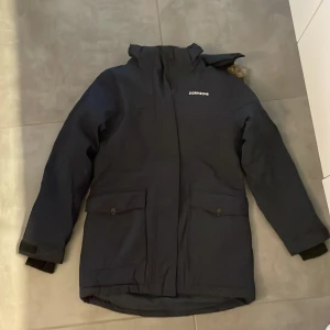 Mörkblå parkas från Didriksons - Mörkblå parkas från Didriksons med huva och fuskpälsdetalj. Jackan är fodrad med mjuk fleece och har två stora fickor framtill, justerbara ärmslut och dragkedja. Perfekt för kalla dagar och helt vattentät. Fin värmande Jacka som går ner över rumpan (storlek 160). 