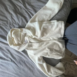 Vit teddyjacka med huva från FILA XS - Mysig vit teddyjacka från FILA i storlek XS. Jackan har huva, dragkedja framtill och två fickor. Tillverkad i fluffigt teddy-material som är perfekt för kyliga dagar. Enkel och stilren design som passar till det mesta.