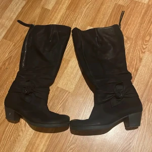 Svarta stövlar med rosettdetalj - Snygga svarta stövlar i mocka med dekorativ rosett vid ankeln och dragkedja på insidan. De har en låg klack och rund tå, vilket gör dem både stilrena och bekväma. Perfekta för dig som vill ha en klassisk look med en twist.