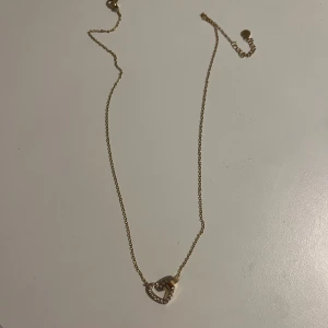 Guldigt halsband med hjärthänge från Edblad  - Supersött halsband i guldton med ett hjärthänge dekorerat med små glittrande stenar. Hjärtat har en öppen design och en rund detalj i guld som sitter fast i kedjan. Perfekt accessoar för att lyfta vilken outfit som helst. Inte använt mycket och är så gott som ny. Pris kan eventuellt diskuteras.