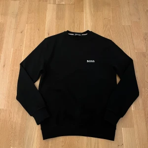 Svart sweatshirt från BOSS - Svart sweatshirt från BOSS med stilren vit logga broderad på bröstet. Tröjan har rund halsringning, långa ärmar och är tillverkad i mjuk bomull. Perfekt för en clean och enkel look.