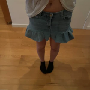 Blå handgjord jeanskjol med volang  - Supersöt blå handgjord jeanskjol från lager 157 i storlek S. Kjolen är kort och har en snygg volangkant nedtill som ger en extra touch. Klassisk femficksmodell med knapp och dragkedja framtill. Perfekt för dig som gillar denim och vill ha något trendigt i garderoben.