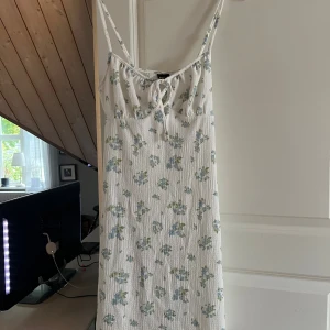 Vit blommig sommarklänning Gina Tricot - Superfin vit kort klänning från Gina Tricot med blått och grönt blommönster. Smala axelband, knytning framtill och lätt strukturerat tyg. Perfekt för varma dagar och ger en söt, avslappnad vibe.