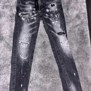 Svarta skinny jeans från Dsquared2 - Säljer ett par svarta skinny jeans från Dsquared2 med slitna detaljer, hål och vit färgstänk. Jeansen har en tight passform och klassisk femficksdesign. Materialet är jeans med stretch för extra komfort. Perfekta för dig som gillar en edgy streetstil.