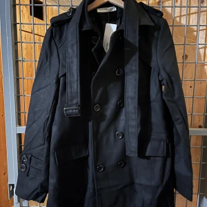 Svart trenchcoat från Msz:Homme - Stilren svart trenchcoat från Msz:Homme i klassisk modell med dubbelknäppning, axelklaffar och bälte i midjan. Jackan har långa ärmar, knappar vid ärmslut och två stora fickor framtill. Perfekt för dig som vill ha en tidlös och snygg look.