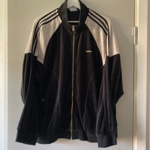 Svart och vit Adidas jacka - Säljer en svart och vit Adidas bomberjacka i mjuk sammet med klassiska tre ränder på axlarna och guld dragkedja. Jackan har lång ärm och broderad Adidas-logga på bröstet. Perfekt för dig som gillar sportig och retro stil. Inte fodrad, mer som en sweatshirt. Uppskattar storlek till L. Säljer för 300 kr + frakt (köpare står för frakt) 💕