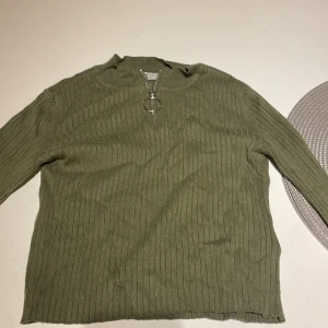 Olivgrön ribbad tröja från H&M - Olivgrön ribbstickad tröja från H&M med lång ärm och rund hals. Tröjan har en dekorativ dragkedja framtill med en stor ring som detalj. Perfekt för dig som gillar en enkel men ändå trendig look.