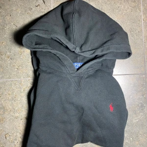 Svart hoodie från Polo Ralph Lauren - Svart hoodie från Polo Ralph Lauren med klassisk röd logga broderad på bröstet. Tröjan har huva, känguruficka och långa ärmar. Tillverkad i mjuk bomullsmix som är perfekt för kyligare dagar. Passar till 150-160 cm lång