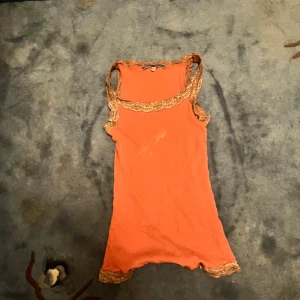 Orange ribbad linnetopp med spets - Snygg orange ribbad linnetopp med breda axelband och dekorativ beige spets runt halsringning, ärmhål och nederkant. Toppen är ärmlös och har en figurnära passform, perfekt för att ge en färgklick till din outfit.
