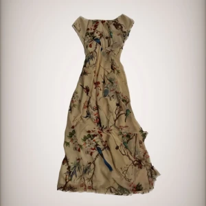 Blommig långklänning med fågelmönster - Säljer en superfin långklänning med ett vackert blommigt mönster och fåglar. Klänningen är i en ljus beige färg och har en romantisk stil. Perfekt för sommarens fester eller en mysig vårdag. Den är ärmlös och har en lös passform som gör den bekväm att bära. Materialet känns lätt och luftigt, vilket gör den idealisk för varmare väder.