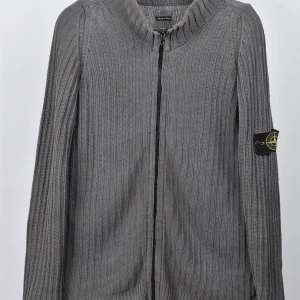 Grå stickad kofta från Stone Island - Säljer en snygg grå stickad kofta från Stone Island. Den har en dragkedja framtill och en ribbad design. Koftan är långärmad och har Stone Island-loggan på vänster ärm. Perfekt för höst och vinter!