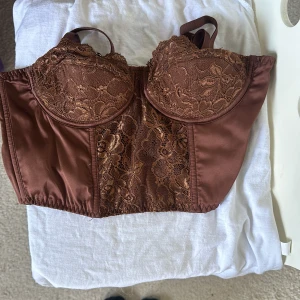 Brun korsett med spetsdetaljer - Säljer en snygg brun korsett från Ann Summers i storlek EU 34. samt ett svart linne från NAKD. Dem har vackra spetsdetaljer och är gjord av satinmaterial. Perfekt för festliga tillfällen eller när du vill känna dig extra fin. Dem har justerbara axelband. Den bruna har  hakknäppning bak för bästa passform. den svarta har en dragkedja på sidan för att få på sig den lätt.🤎🖤