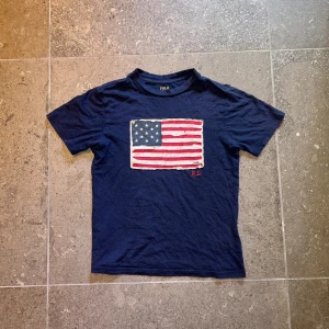 Mörkblå t-shirt från Polo Ralph Lauren - Säljer en mörkblå t-shirt från Polo Ralph Lauren med en cool amerikansk flagga på framsidan. Flaggan är lite sliten för en vintage look och har initialerna R.L. broderade i rött. T-shirten är i bomull och har korta ärmar. Perfekt för en casual look!