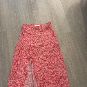 Röd blommig långkjol - Säljer en superfin röd långkjol med små vita blommor. Kjolen har en slits framtill.. Materialet känns lätt och luftigt, vilket gör den bekväm att bära hela dagen. Upplever den mindre i storlek. 