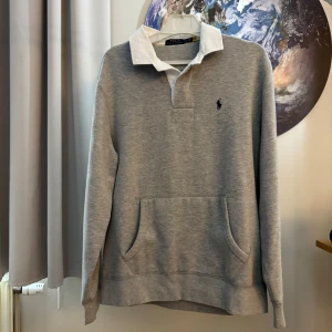 Grå tröja från Ralph Lauren - Säljer en snygg grå tröja från Ralph Lauren. Den har en vit krage och en liten broderad logga på bröstet. Tröjan har långa ärmar och en praktisk ficka på magen. Perfekt för en casual look! Oanvänd!