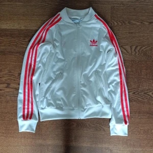 Vit Adidas jacka med röda ränder - Säljer en snygg vit Adidas zip-up med röda ränder längs ärmarna och Adidas logga på bröstet och ryggen. Jackan har dragkedja framtill och två fickor med dragkedja. Perfekt för en sportig look eller vardagsstil. Passar bra till både träning och fritid.