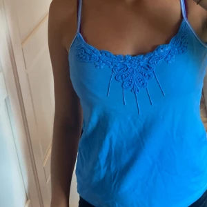 Blå linnetopp med spetsdetaljer - Säljer en superfin blå linnetopp med smala axelband och vacker spetsdetalj vid halsringningen. Perfekt för sommaren eller en festlig kväll. Toppen är i bra skick och väldigt bekväm att ha på sig. Passar perfekt till både jeans och kjol!