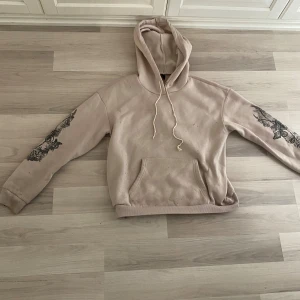 Beige hoodie från Romwe - Säljer en snygg beige hoodie från Romwe i storlek M. Den har en cool svart tryckdesign på ärmarna och ryggen. Hoodien är perfekt för höst och vinter, och den är superbekväm att ha på sig. Den har en känguruficka fram och en justerbar huva med dragsko. Perfekt för en casual look eller för att mysa hemma!