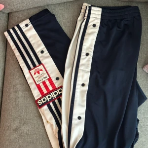 Svarta Adidas byxor med vita ränder - Säljer ett par Adidas popper pants i storlek M. Adibreak. De är gjorda av ett syntetmaterial och har en elastisk midja för extra komfort. Perfekta för träning eller en avslappnad dag. Köp direkt eller ge prisförslag!