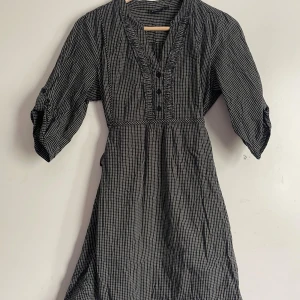 Svart och vit rutig klänning från Lindex - Säljer en superfin svart och vit rutig klänning från Lindex i storlek 42. Klänningen har trekvartsärmar och en knytning i midjan för en fin passform. Den har också knappar framtill och en v-ringad hals. Perfekt för både vardag och fest!