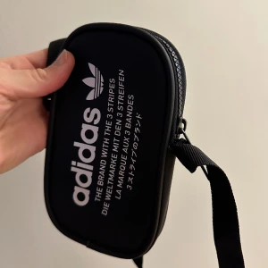 Väska Adidas - Liten väska från Adidas. Får plats med mobiltelefon och lite andra små grejer. Axelrem följer med. Nästan aldrig använd.