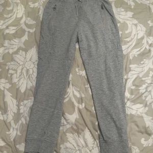 Grå mjukisbyxor - Ett par grå mjukisbyxor från Urban Future Young Learners i storlek 150. Byxorna har elastisk midja med dragsko, två sidofickor med dragkedja och en bakficka. De har ribbade muddar vid bensluten och är tillverkade i ett mjukt bomullsmaterial. Byxorna har några fläckar.