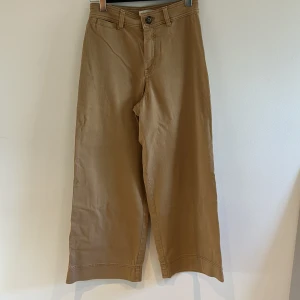 Beige byxor från H&M - Ett par högmidjade beige byxor från H&M i storlek 34. Byxorna har en rak vid passform och är tillverkade i ett bomullsmaterial. Modellen är wide leg.