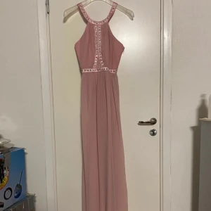 Rosa långklänning med pärldetaljer - En elegant rosa långklänning med pärldetaljer runt halsen, midjan och längs framsidan. Klänningen har en dragkedja på sidan och är ärmlös med en öppen ryggdesign.💕