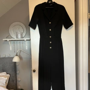 Svart jumpsuit  - En festlig svart jumpsuit med v-ringning, knappar och uppvikt kant nedtill.  Extra knapp finns, i fint skick! 