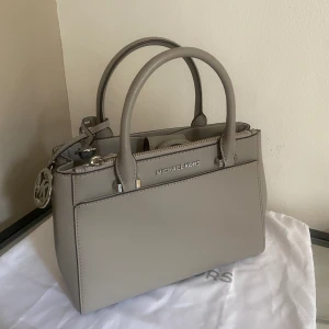 Michael Kors - Hej!  Jag säljer min super fina Michael Kors väska som är i helt nytt skick.  Väskan är i färgen Pearl Grey & modellen heter Gibson-Satchel.  Tillsammans med väskan får du med den vita Michael Kors påsen på bilden, oanvänt axelband samt original tag