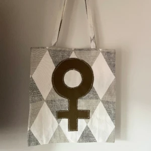 Tygkasse, tygväska, tygpåse  - Handsydd tygkasse med motivet ”feminist”.   Tillverkad i 100% återvunna textilier.   Mått: 32x35 cm  Hämtas på Södermalm alternativt skickas med post. 