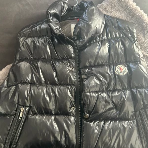 Moncler väst  - Klassisk elegans möter modern detaljering. Denna väst ger en sofistikerad touch till din outfit samtidigt som den håller dig varm och bekväm. En tidlös Moncler-kreation för alla tillfällen.