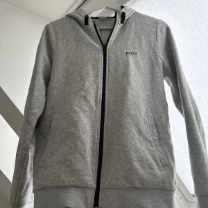 Hugo Boss tröja - en riktigt snygg grå hugo boss zip tröja i en grå fet färg men ett snyggt tryck på ryggen , skick 10/10 aldeig använd pga var förliten , hör av er vid frågor&bilder🤩🤩