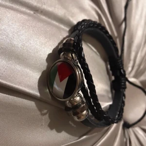 Svart flätat armband med flaggdetalj - Ett svart flätat Palestina flagga armband med en rund metallberlock som har en flaggdetalj i rött, vitt, svart och grönt. Armbandet har justerbar knytning för att passa olika storlekar.