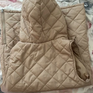 Beige quiltad väst med huva från 157 - En beige quiltad väst med huva från märket 157. Väst har en dragkedja framtill och två fickor med dragkedja. Den är i storlek 150 och är lämplig för kyligare väder.