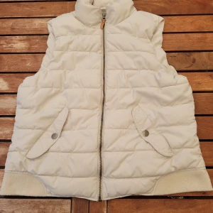 Vit dunväst från L.O.G.G. - En vit dunväst från L.O.G.G. med dragkedja och två fickor framtill. Västen har en hög krage och knappar på fickorna.