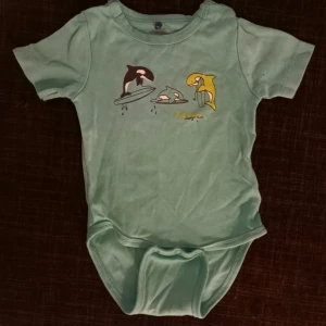 Kortärmad body strl. 6-9 mån - Grönaktig kortärmad body med späckhuggare i storlek 68 (6-9 mån) från Mee too. I fint begagnat skick, inga hål men en dov fläck ovanför den svarta späckhuggaren. Unisex  Tvättad i parfymfritt tvättmedel och utan sköljmedel.