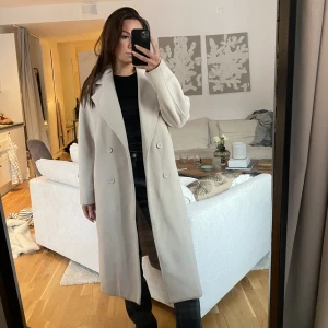Beige lång kappa - Beige lång kappa. Liten slits på båda sidor, knappar i fram & två fickor.