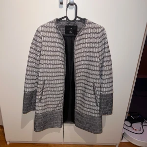 Svartvit kappa från H&M - En elegant svartvit kappa från H&M med ett mönster som liknar små rutor. Kappan har långa ärmar och en rak passform. Den är fodrad på insidan för extra komfort.