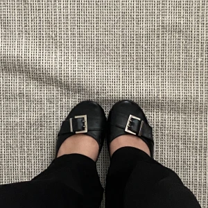 Svarta ballerinaskor med spänne - Ett par svarta ballerinaskor med ett dekorativt spänne på ovansidan. Skorna har en rund tå och en låg klack. Från märket Roberto Durville, vintage Strolek 37