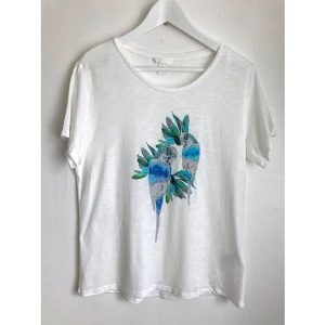 Vit t-shirt med turkosa papegojor, stl M, pärlor, tryck, tropisk, fågel - Vit t-shirt med papegojtryck i grönt och blått samt transparenta pärlor. Mått i cm: bröstvidd 48; axelbredd 41; längd fram från axeln 62; ärmlängd 18.