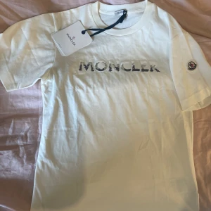 Moncler t-shirt  - En vit t-shirt med tryck där det står moncler med glittrig text. Supersöt till våren/sommaren. Om ni vill ha mer bilder eller har frågor skriv till mig❤️Pris kan diskuteras. Nypris 3100 aldrig använd❤️