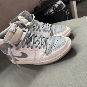 Jordan 1 höga - Hej Air Jordan 1 i klassiskt vitt och grått, storlek 38,5 EU. Dessa tidlösa sneakers har använts varsamt och är i bra skick med några lätta veck (creases) som bidrar till karaktären. De är perfekta för både samlare och modeentusiaster som söker en au