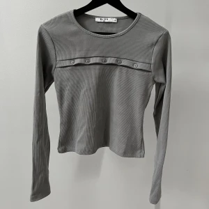 Grå långärmad med knappar NA-KD - En grå långärmad tröja med knappar på från NA-KD x Chloé Schuterman. Använd en gång. Knapparna går att knäppa eller ha öppna. Nypris 399kr.