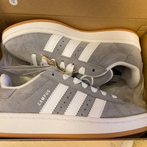 Nya Adidas Campus 00s -storlek 38 - Adidas Originals Campus 00s j storlek: 38.  Färg: Grå (Grey Three / Cloud White / Cloud White) Ingår både vita och gråa skosnören.  Nya och oanvända. Ligger kvar i kartong.  Köpta i Sverige och inte från någon massbeställning utomlands. Äkta Campus. 