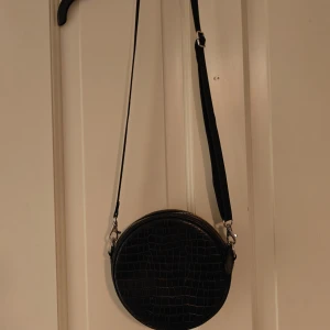 Svart rund axelbandsväska  - Köpt secondhand men i fint skick, knappt tecken på användning Justerbart och avtagbart axelband. Har ett litet extra fack på insidan