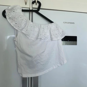 Vit off-shoulder topp - En vit off-shoulder topp med volangdetaljer. Toppen har ett broderat mönster på volangen och är ärmlös.