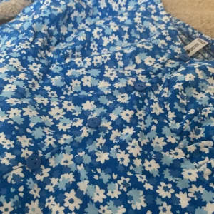 Blommig topp från FB Sister - En blå blommig topp från FB Sister med vita och ljusblå blommor. Toppen har långa ärmar och knappar framtill. Den är tillverkad i ett elastiskt material för en bekväm passform.