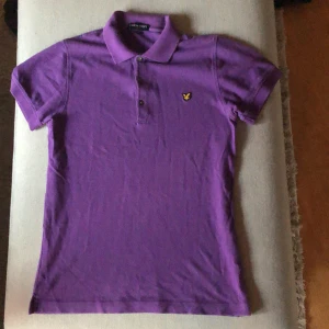 Lila pikétröja från Lyle & Scott - En stilren lila pikétröja från Lyle & Scott med korta ärmar och en klassisk krage. Tröjan har tre knappar framtill och ett broderat gult Lyle & Scott märke framtill